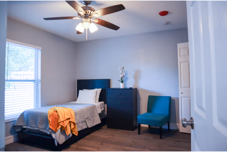 A simple bedroom with a single bed covered with a gray quilt and an orange blanket draped over the foot. There is a black dresser with a vase of flowers on top, a teal upholstered chair, a window with blinds, and a ceiling fan with lights.