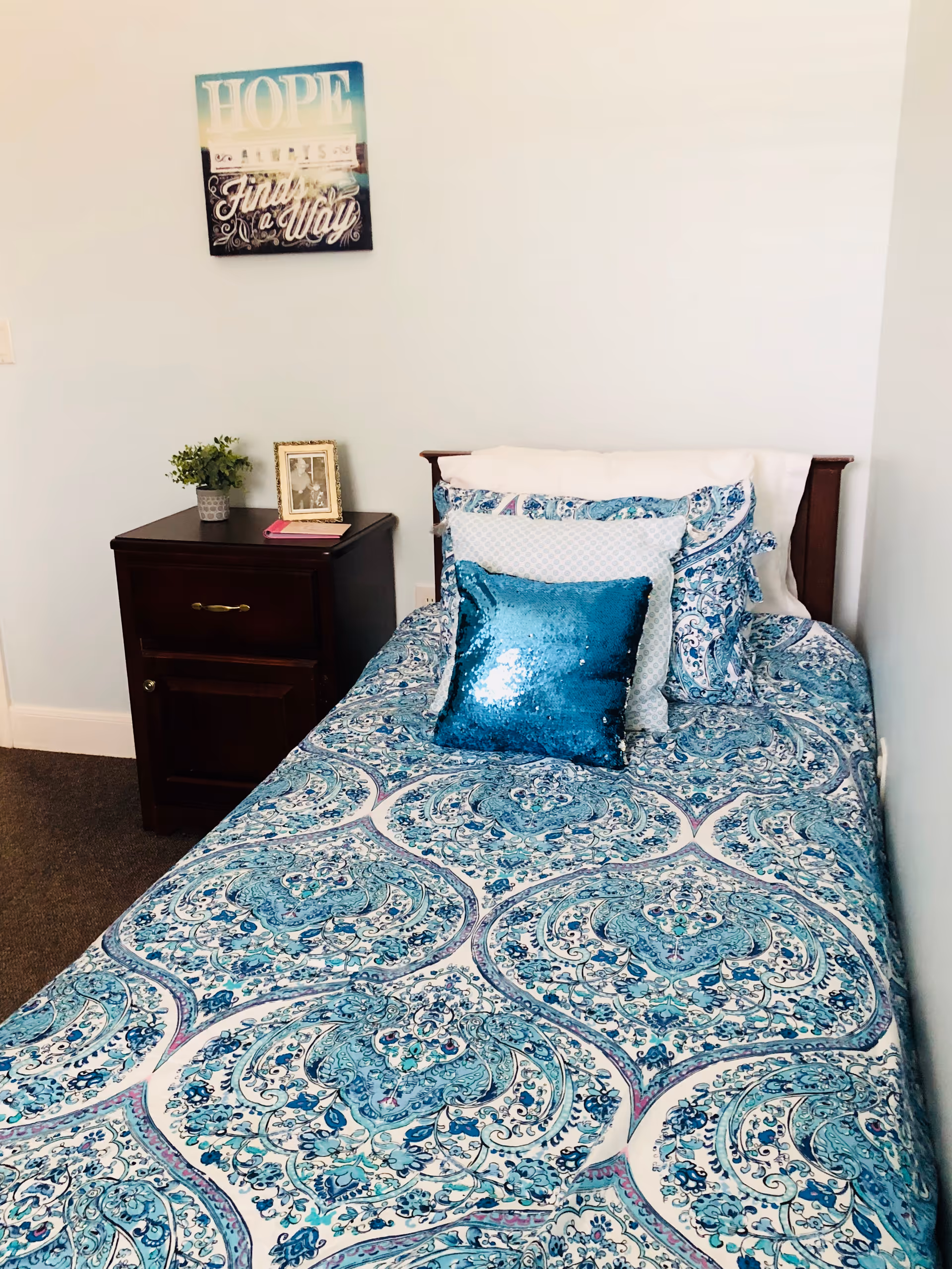 Single bed with blue patterned bedding and a teal sequined pillow next to a dark wood nightstand and a 'HOPE' wall art above.
