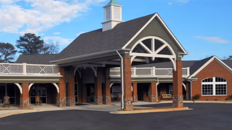 Front entrance of a brick senior living building with a covered porte-cochère and second-story balcony.