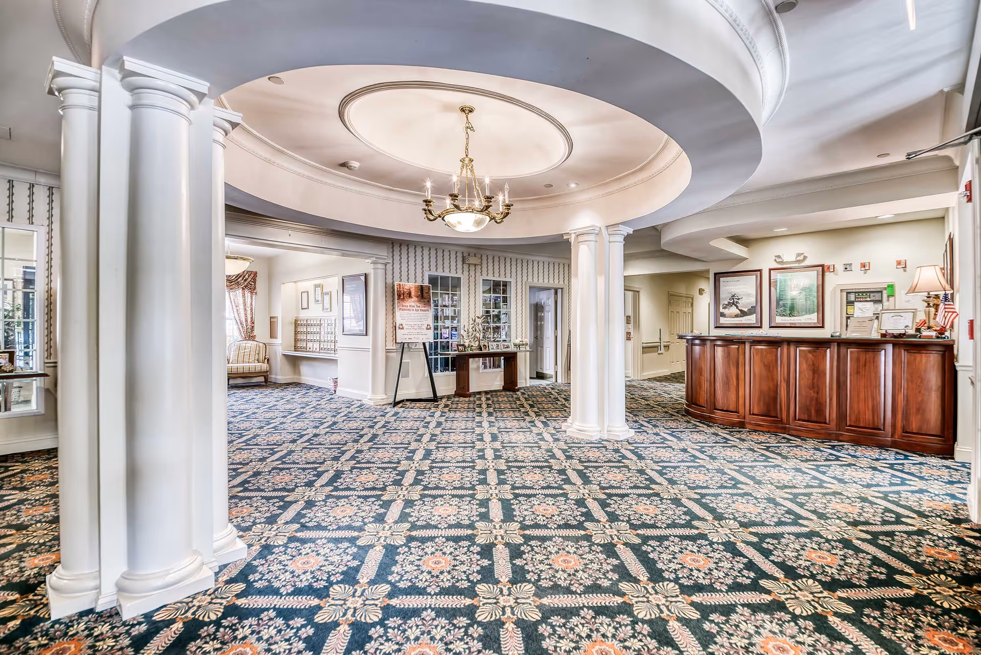 Spacious senior living facility lobby with patterned carpet, white columns, a circular ceiling design with a chandelier, a wooden reception desk, and framed artwork on the walls.
