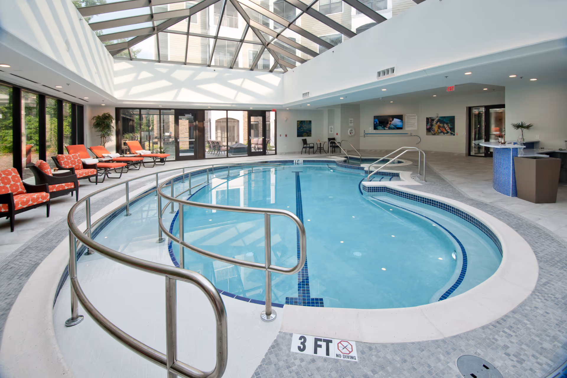 Indoor swimming pool area with a glass ceiling allowing natural light to fill the space. The pool has stainless steel handrails and a shallow entry marked 3 feet deep with a no diving sign. Around the pool are orange cushioned lounge chairs and armchairs, with some plants and tables. The area has large windows and doors leading outside, and a small bar or reception desk is visible on the right side.
