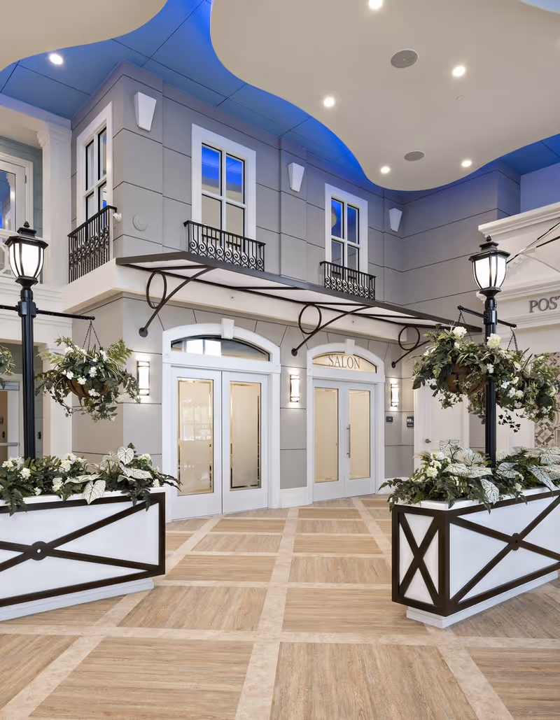 Interior view of a senior living facility hallway with a salon entrance. The area features a high ceiling with blue accent lighting, decorative black metal railings on upper windows, hanging plants, and classic-style lamp posts. The floor has a light wood pattern, and the walls are painted in soft gray tones.