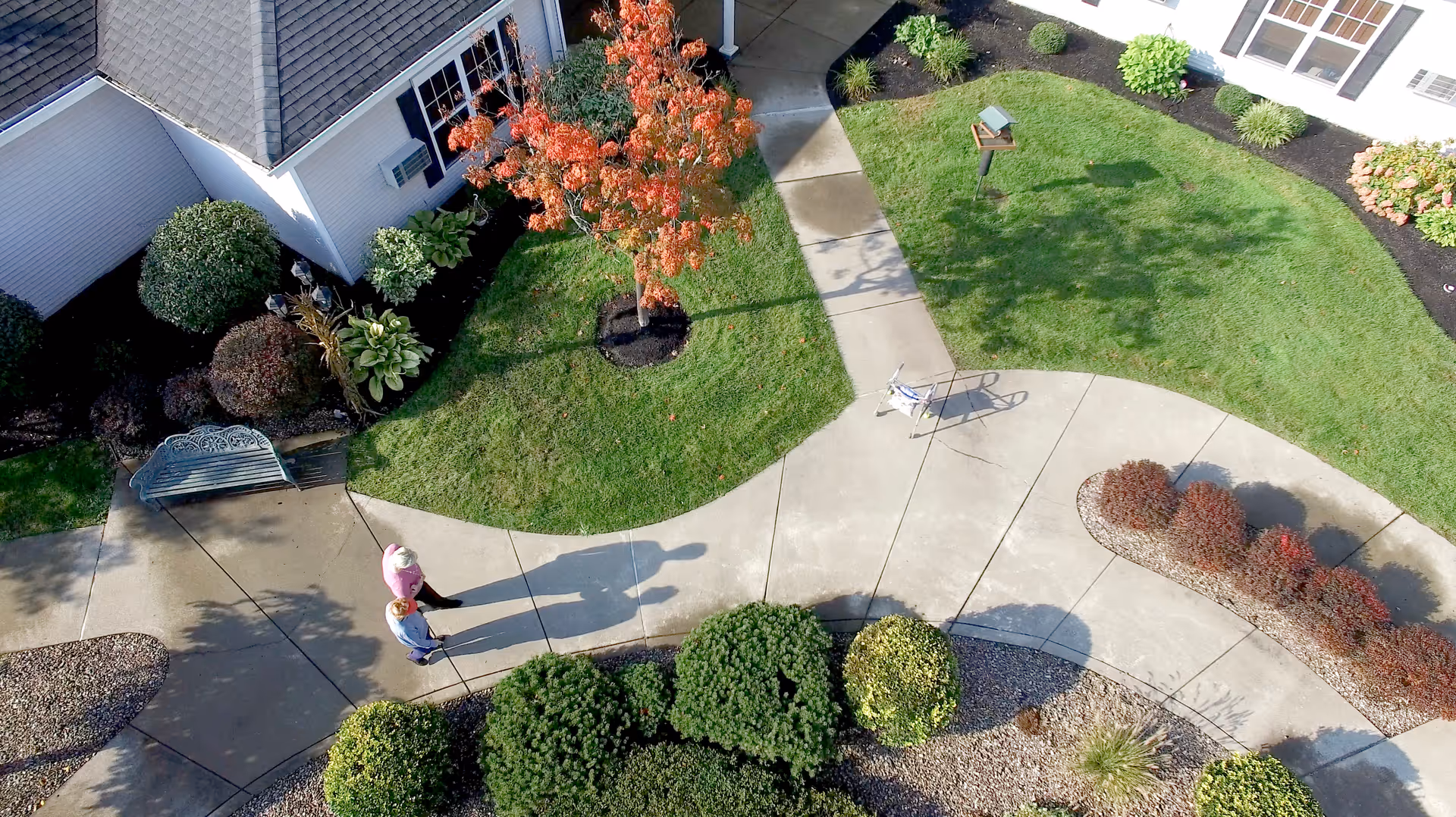 Aerial view of a landscaped courtyard with curved concrete paths, shrubs, a bench, and two people walking.