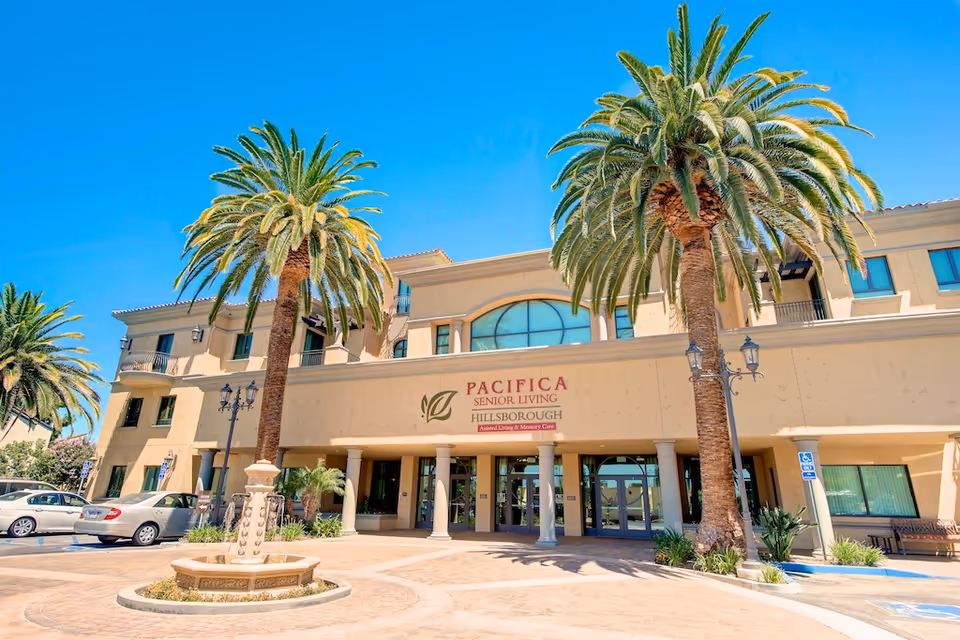 Three-story senior living building entrance with tall palm trees, a fountain, and parked cars under a clear blue sky.