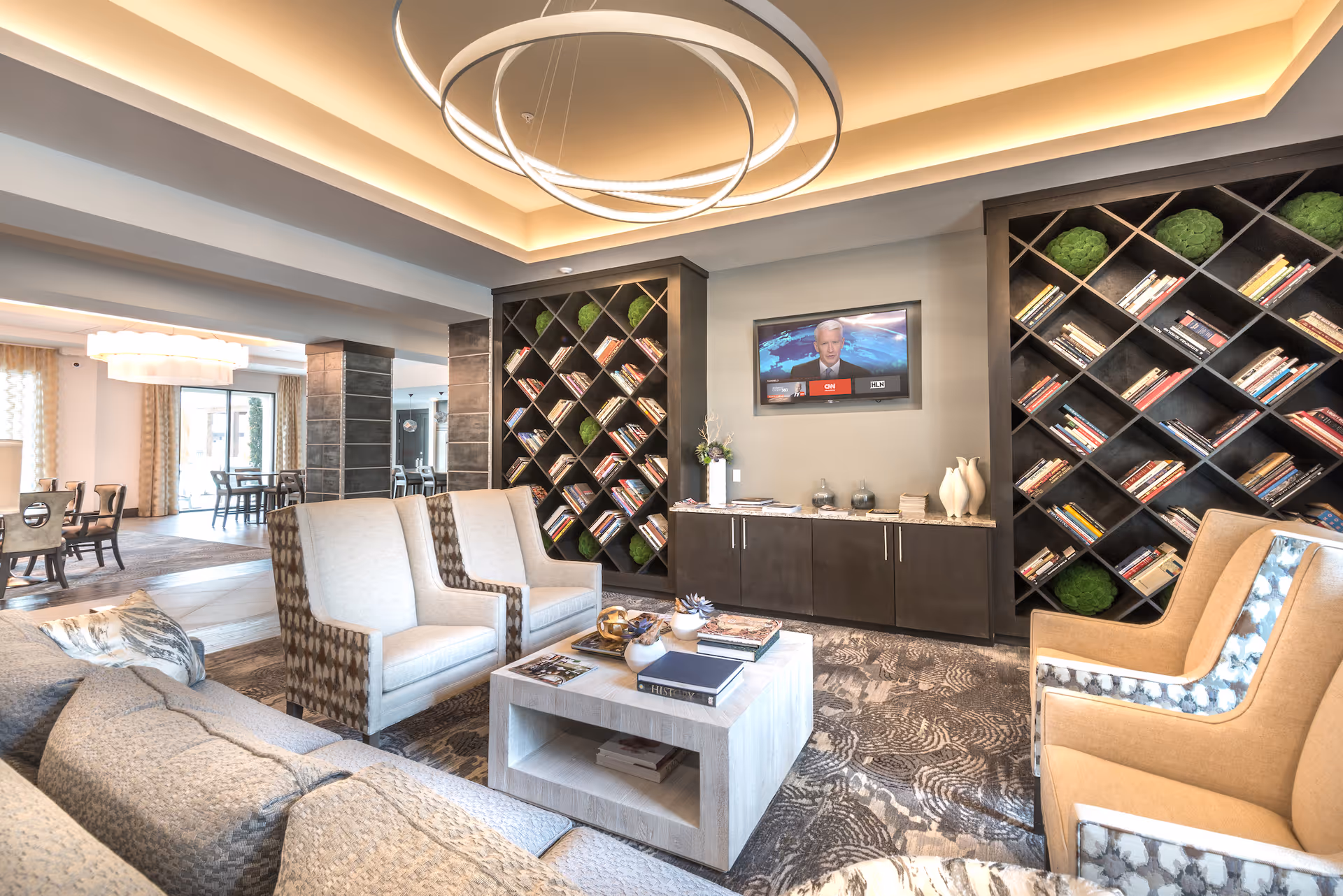 Spacious modern lounge with armchairs and sofas arranged around a coffee table, diagonal bookshelves, a wall-mounted TV, and a circular chandelier.