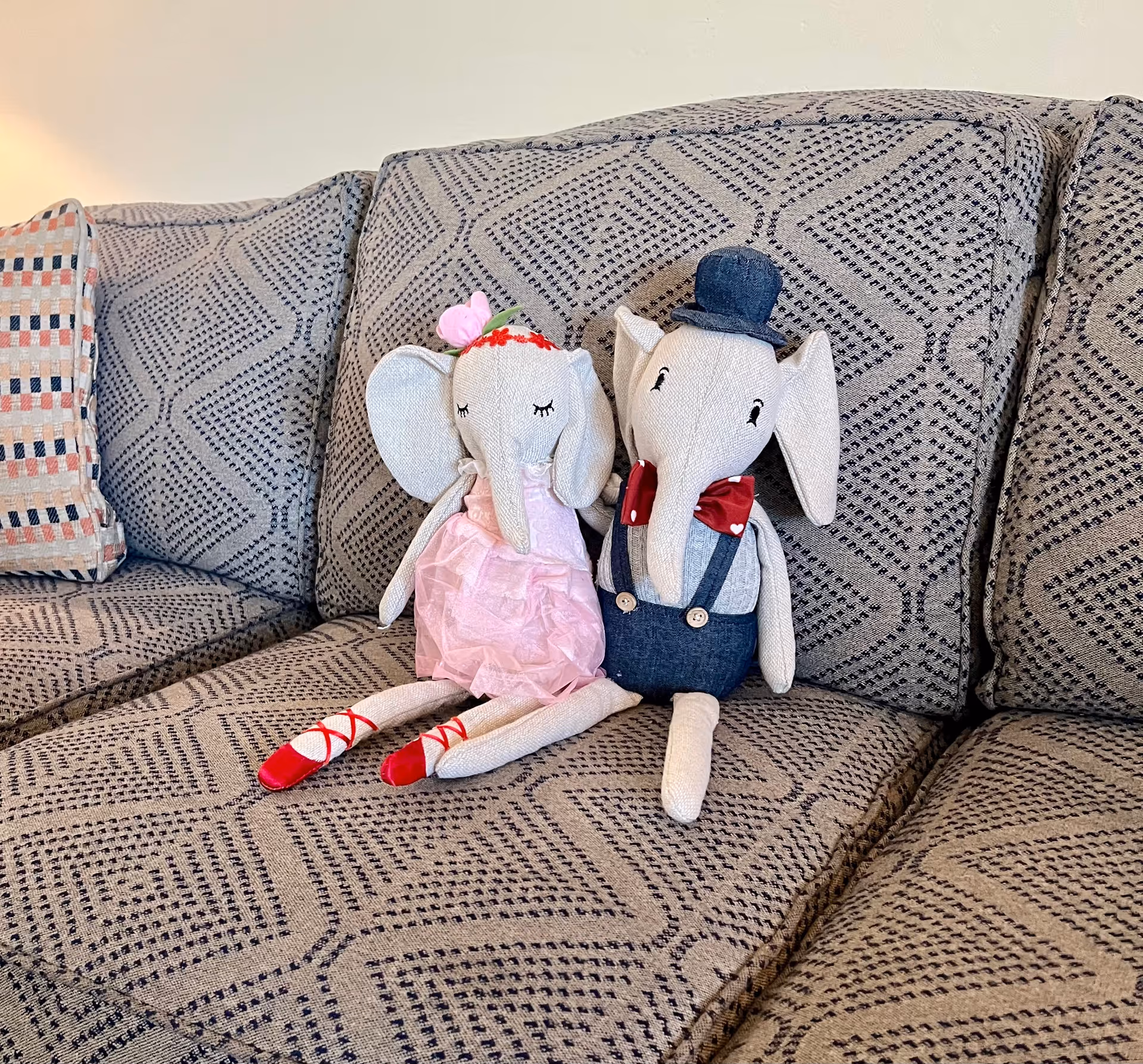 Two stuffed elephant dolls sitting on a patterned gray couch. One elephant is dressed in a pink dress with red ballet shoes and a flower headband, while the other is wearing a blue outfit with suspenders, a red bow tie, and a blue hat.