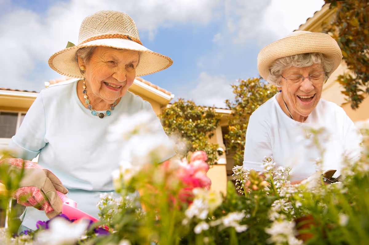 Two elderly women wearing sun hats and gardening gloves are happily tending to flowers in a garden outside a residential building under a partly cloudy sky.