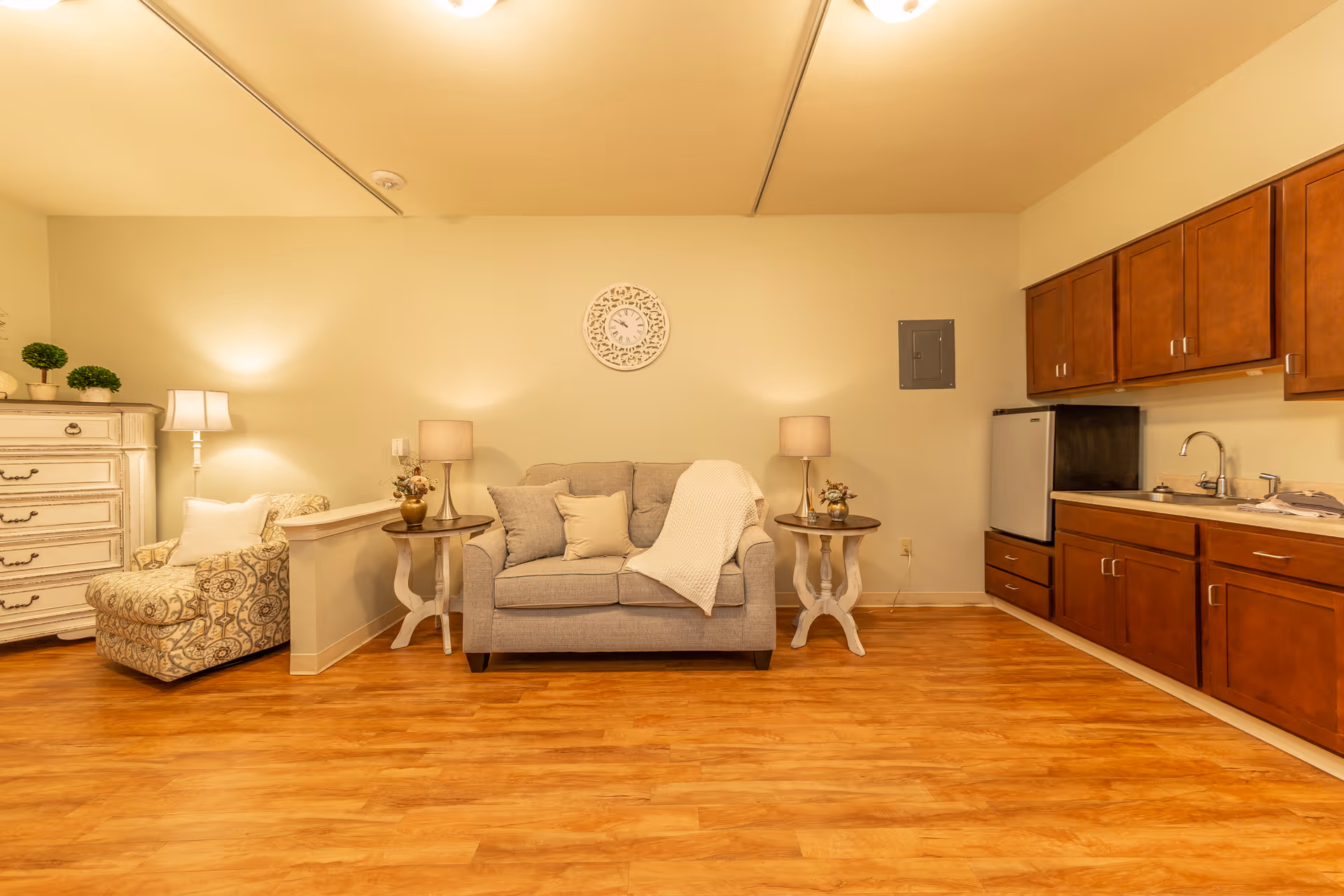 Cozy common room with a gray loveseat flanked by side tables and lamps, a patterned armchair, and a small kitchenette with wooden cabinets.