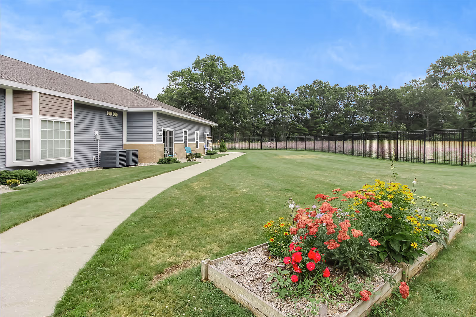 Exterior view of a single-story assisted living building with a curved concrete walkway, manicured lawn, fenced yard, and a raised flower bed of colorful blooms.