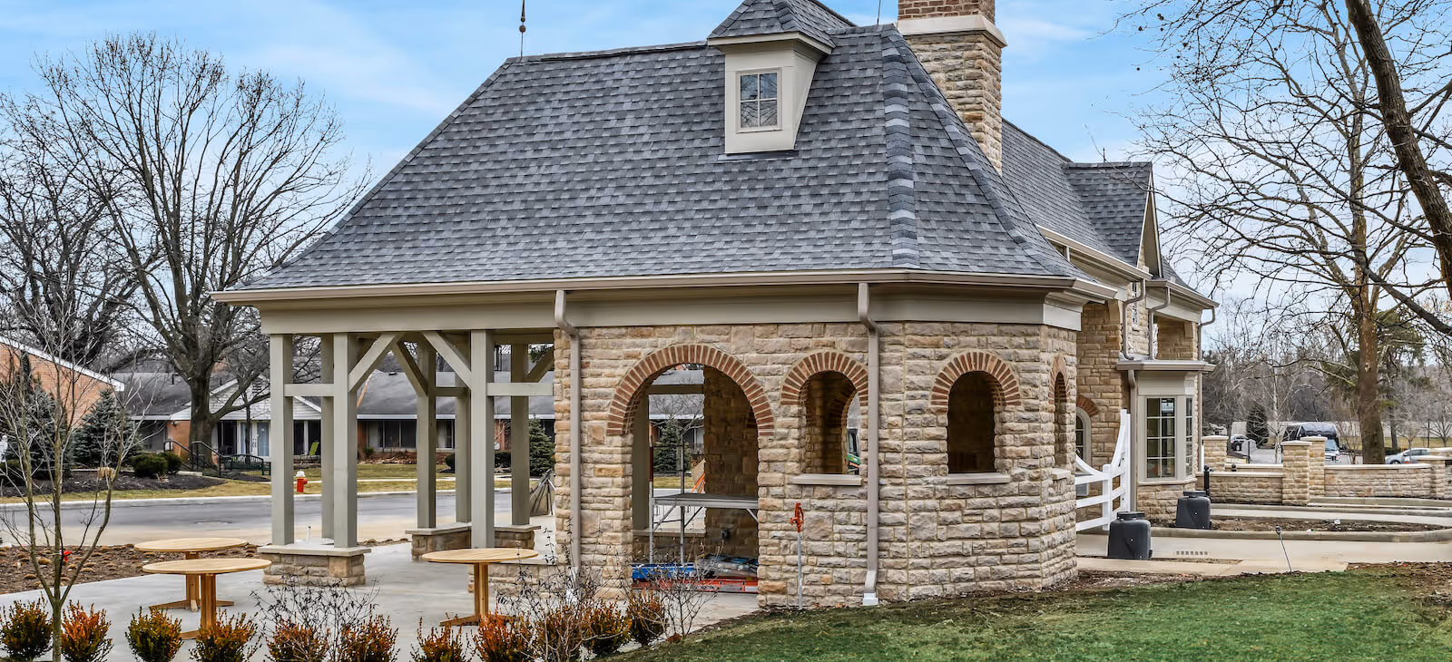 Stone pavilion with arched openings and a shingled roof in an outdoor area of a senior living facility. There are round wooden tables with benches nearby, leafless trees, and a paved walkway surrounding the pavilion. Other buildings and parked cars are visible in the background under a partly cloudy sky.
