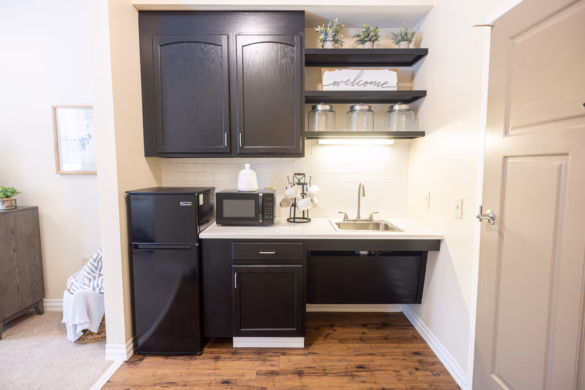 A small kitchenette with black cabinets, a black mini refrigerator, a microwave, a sink, and open shelves holding glass jars and small potted plants. A 'welcome' sign is displayed on the top shelf. The floor is wooden, and there is a door to the right.