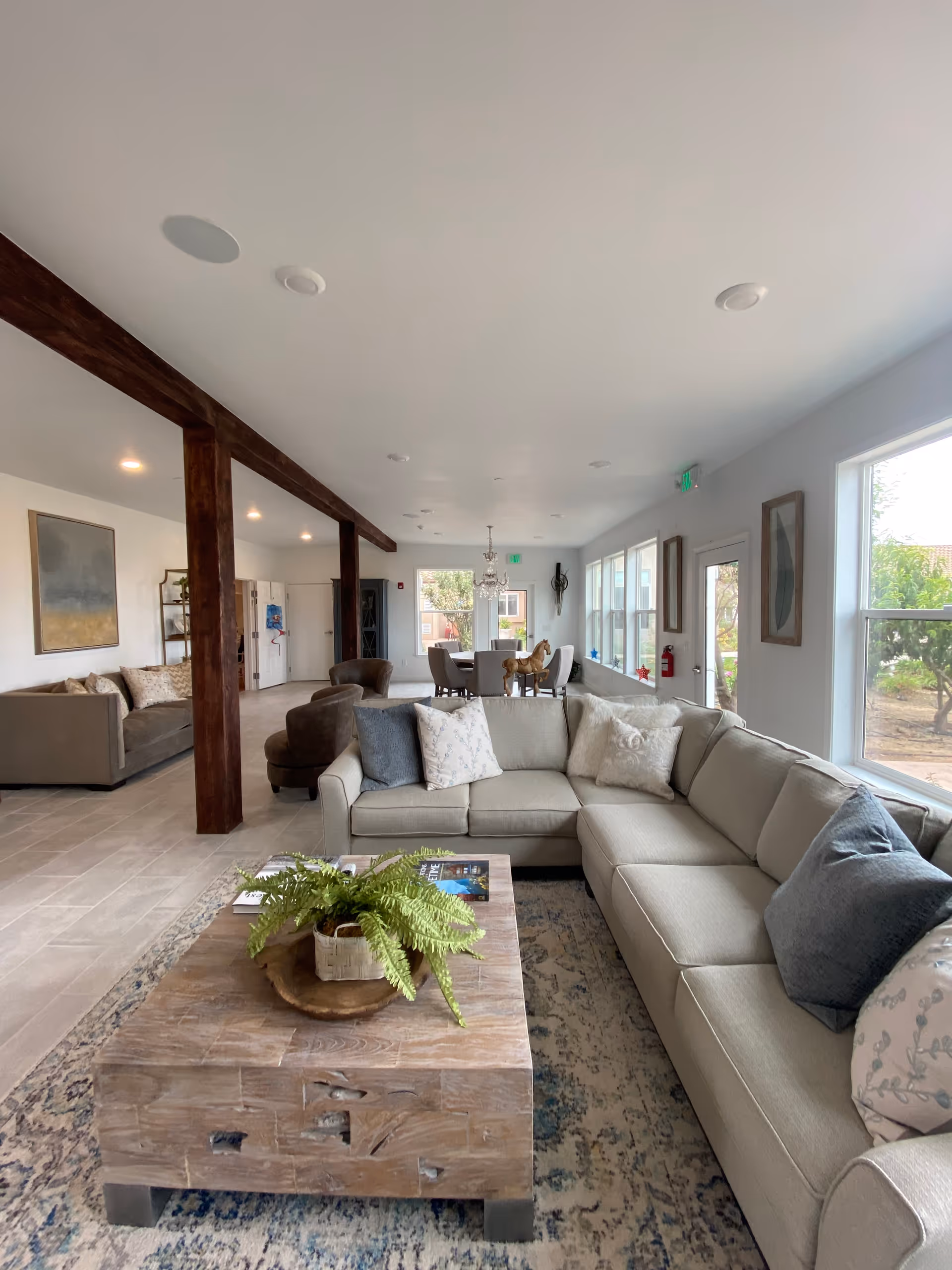 A spacious and bright living room area with a large beige sectional sofa adorned with various pillows. In front of the sofa is a rustic wooden coffee table with a green potted plant and some magazines. The room has large windows on one side letting in natural light, and there are wooden beams on the ceiling. In the background, there is a dining area with a chandelier and chairs, and another seating area with a couch and armchairs.
