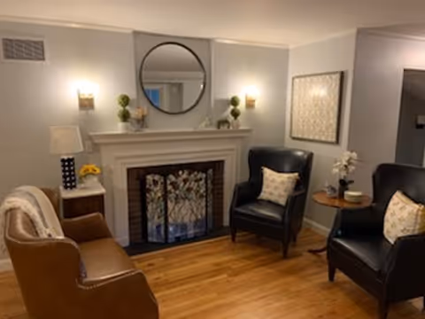 Cozy living room with a white fireplace, round mirror, two black armchairs, a brown leather chair, and hardwood floors.