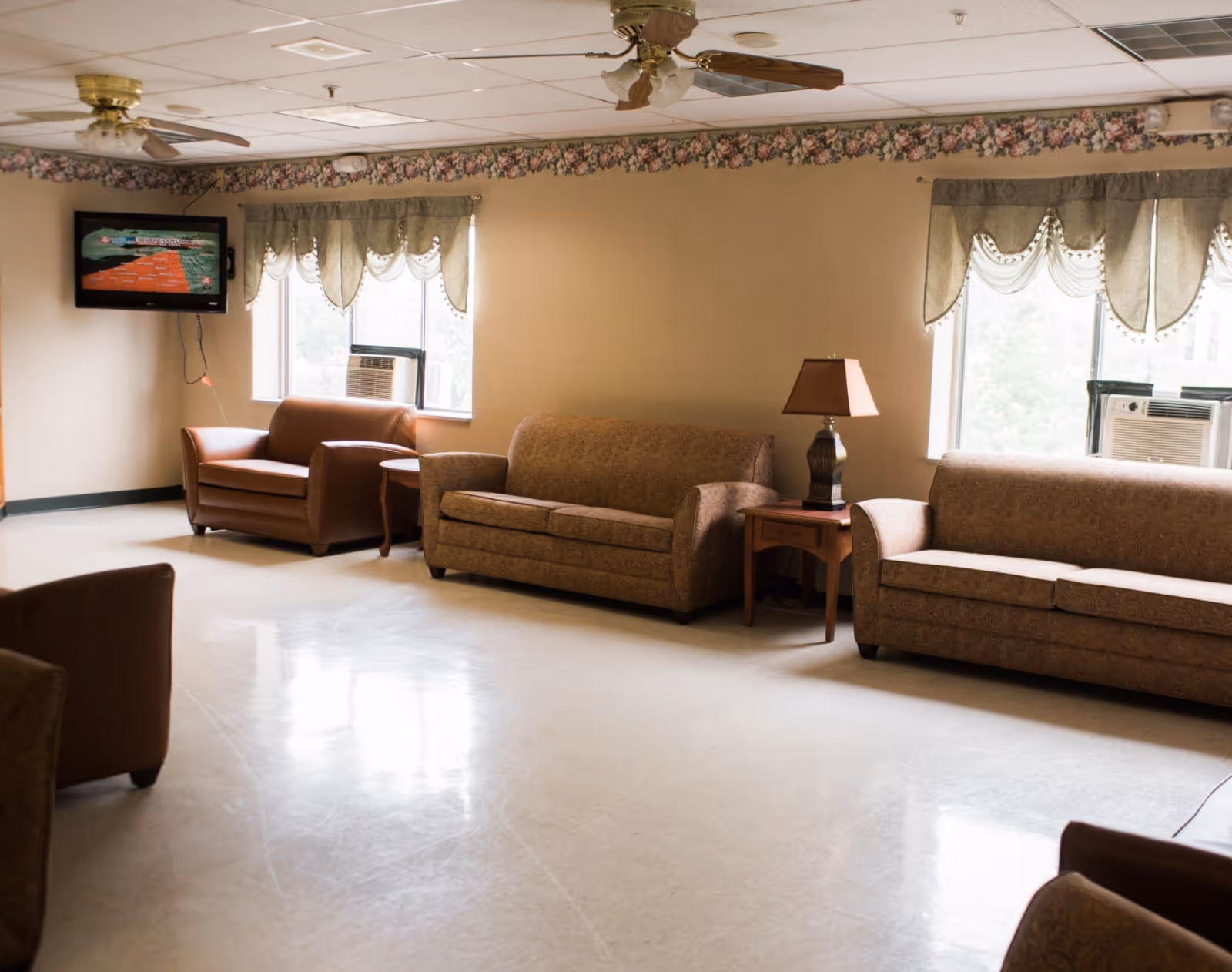 A senior living facility common room with sofas, armchairs, side tables and lamps, a wall-mounted TV, windows with valances, and ceiling fans.