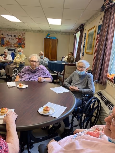A group of elderly individuals sitting around tables in a room, some in wheelchairs, with cupcakes on plates in front of them. The room has beige walls, a window with curtains, and a cabinet in the background. A fall-themed decoration is visible on the wall.