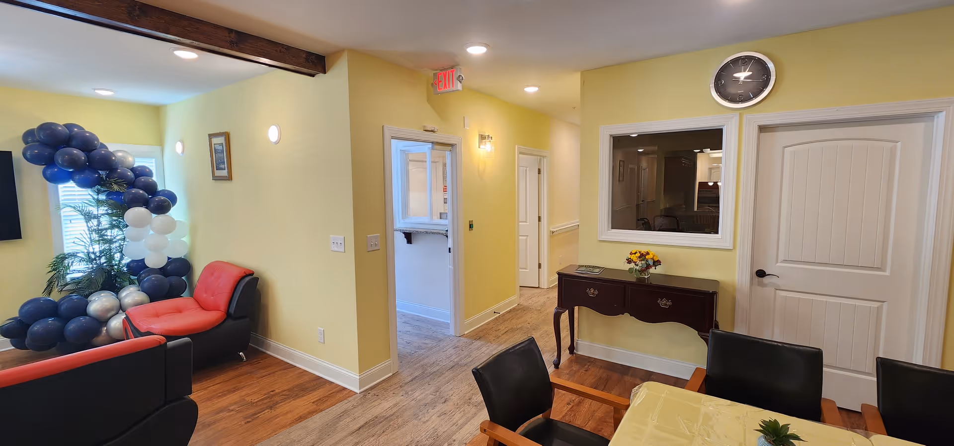 Interior view of a senior care facility room with yellow walls and wooden flooring. The room features a red and black cushioned chair, a table with black chairs around it, a small wooden console table with a flower arrangement, a clock on the wall, and a window looking into another room. There is a balloon decoration with dark blue, white, and silver balloons near a window with blinds.