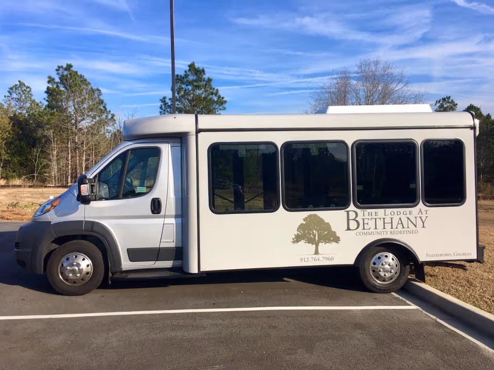 A white shuttle bus parked in a parking lot with trees and a blue sky in the background. The bus has the logo and text for The Lodge At Bethany, including a tree graphic and contact information.