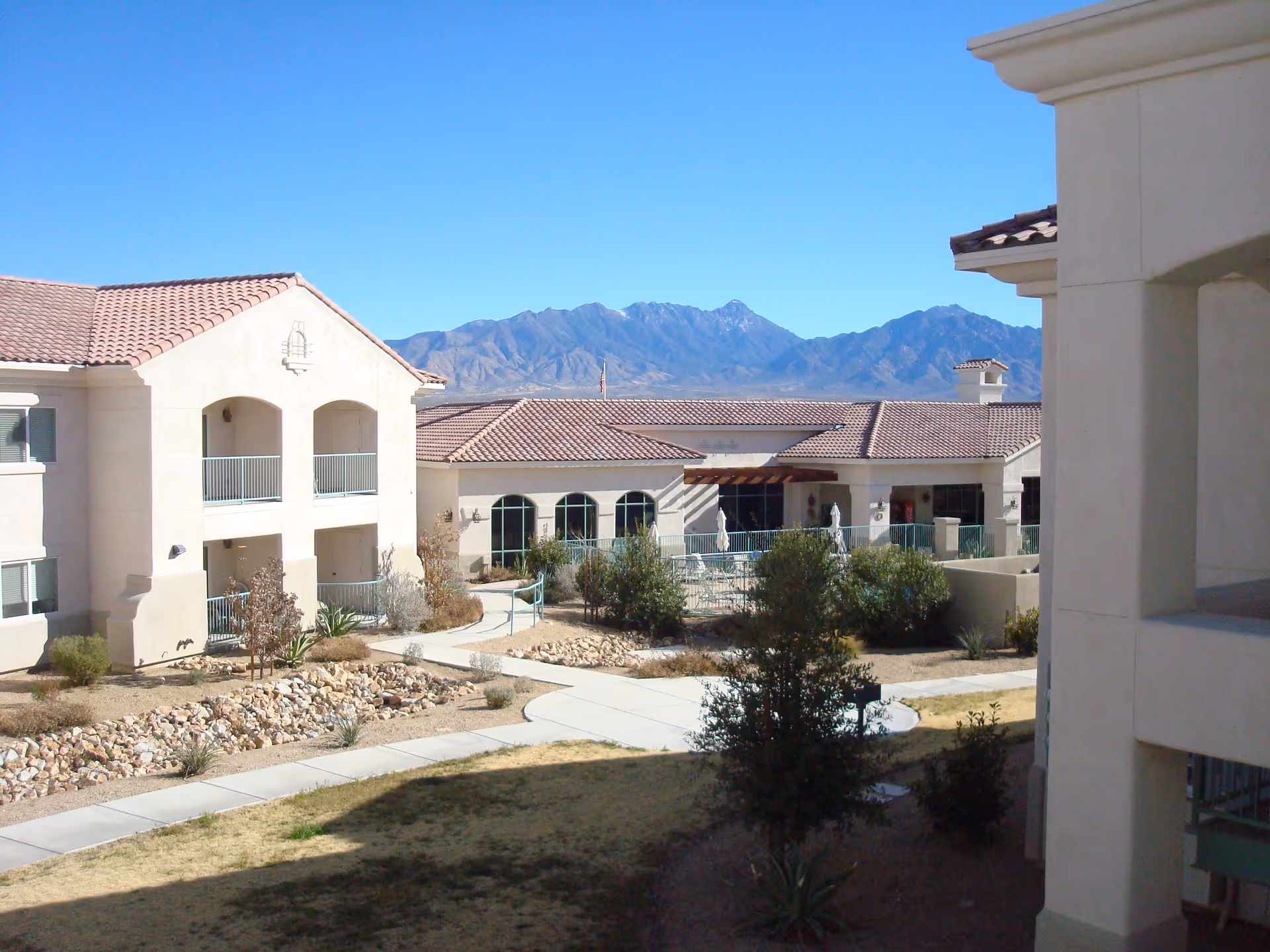 View of a senior living facility courtyard with beige stucco buildings featuring red tile roofs, balconies, and a paved walkway. There are some bushes and small trees in the landscaped area, and mountains are visible in the background under a clear blue sky.