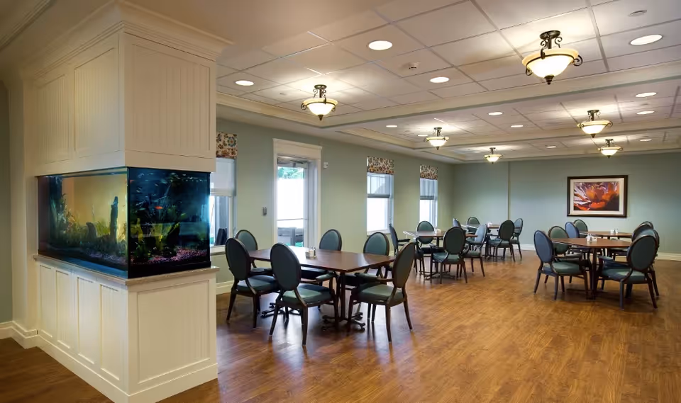 A spacious dining room with several round and rectangular tables surrounded by green cushioned chairs. The room features wood flooring, soft green walls, and multiple windows with floral valances allowing natural light. A large built-in aquarium is visible on the left side, and a framed floral artwork hangs on the far wall. Ceiling lights provide additional illumination.