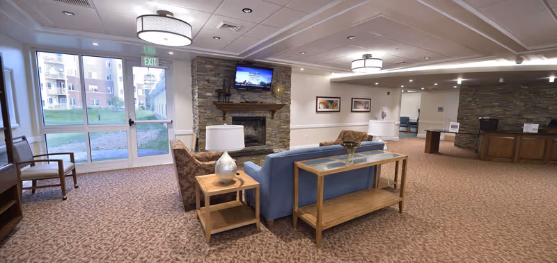 A senior living facility common area with sofas and chairs arranged around a stone fireplace and wall-mounted TV, a reception desk to the right, and glass exit doors to the left.