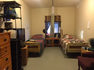 A small room with two single beds separated by a wooden nightstand with a lamp and a flower vase. Each bed has a colorful quilt. There is a window with curtains behind the nightstand. On the left side, there is a wooden dresser and a black metal shelving unit with various items. On the right side, there is a maroon armchair and a small table with a plant and other items. The room has beige walls and a light carpeted floor.