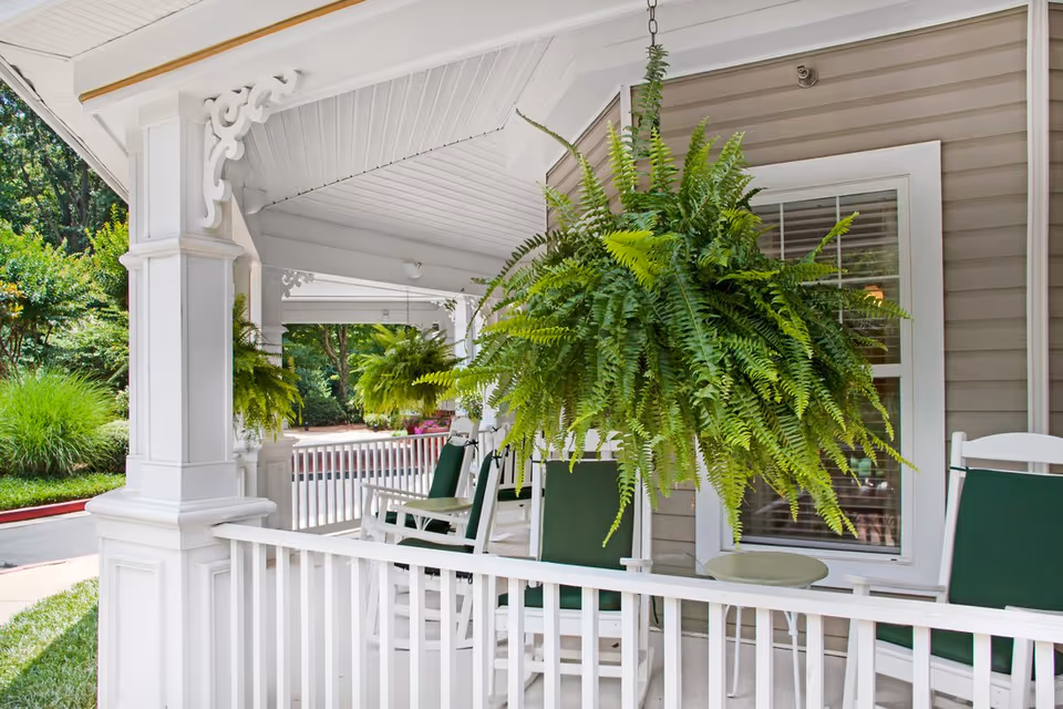 Front porch with white railing, green-cushioned rocking chairs and large hanging fern plants.