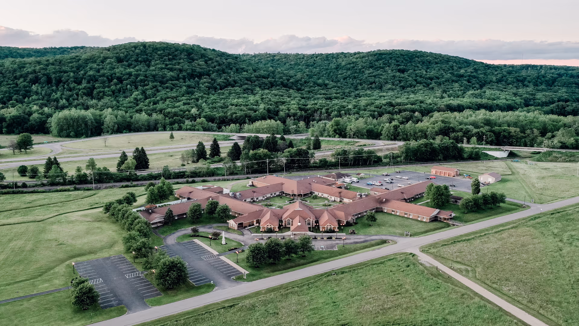 Aerial view of a sprawling single-story senior living campus with red roofs, parking lots, lawns, and wooded hills in the background.