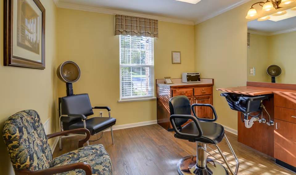 Small salon-style room with a barber chair, hooded hair dryer, sink station, and upholstered waiting chairs by a window.
