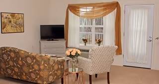A cozy living room with a patterned brown sofa and a white armchair with black dots facing a small white dresser with a flat-screen TV on top. A window with sheer white curtains and a brown swag valance is behind the armchair, and a door with a sheer curtain is to the right. A small round wooden table with a vase of pink flowers is between the sofa and armchair. A framed floral artwork hangs on the wall above the sofa.
