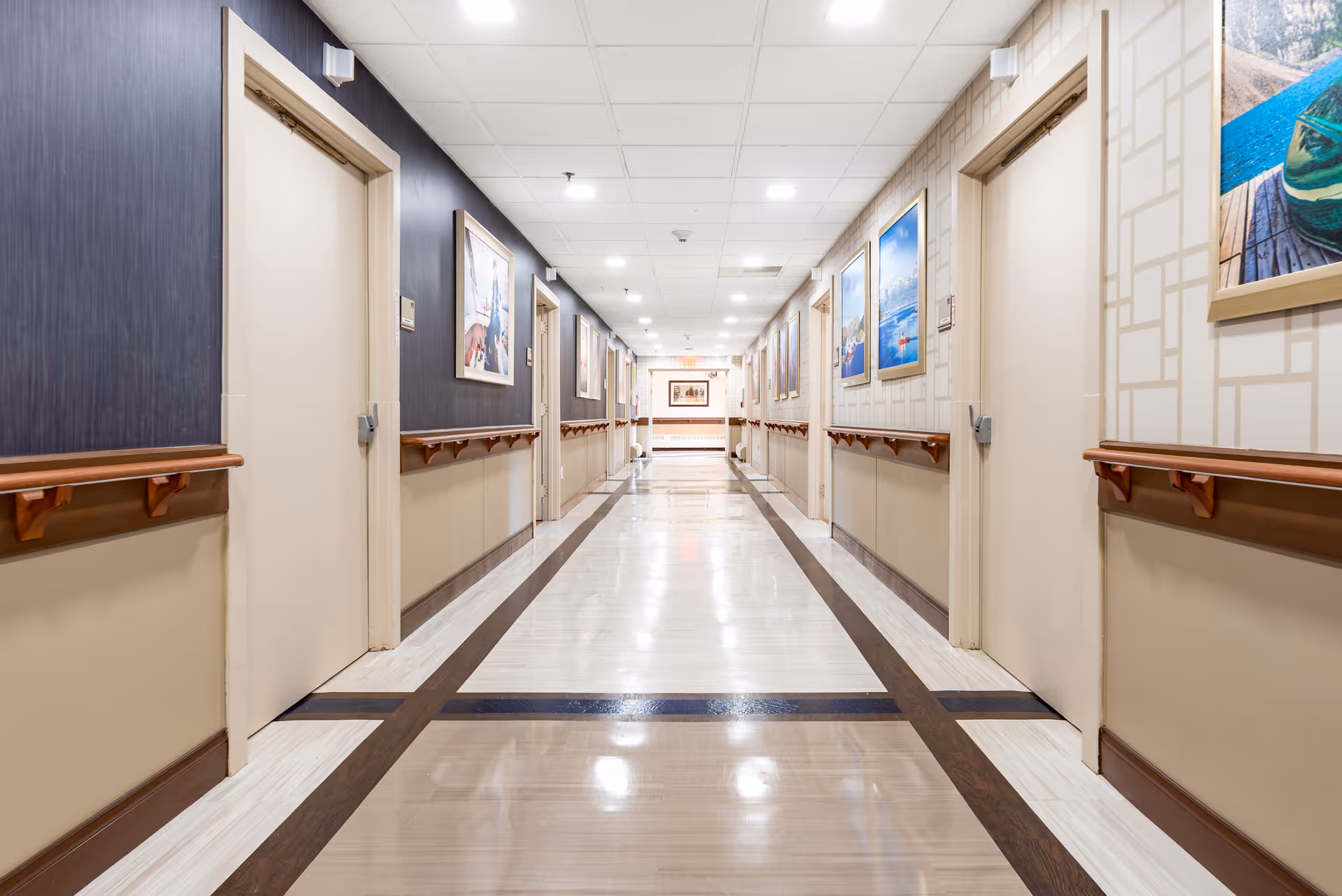 A long, well-lit hallway in a senior living facility with closed doors on both sides, handrails along the walls, and framed pictures hanging above the handrails. The floor has a polished tile pattern with dark borders.