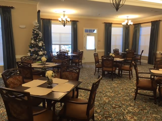 A dining room with several tables and chairs arranged neatly. Each table has a small floral centerpiece. There is a decorated Christmas tree in the corner near windows with blue curtains. The room has warm lighting from ceiling chandeliers and a door with a window at the far end.