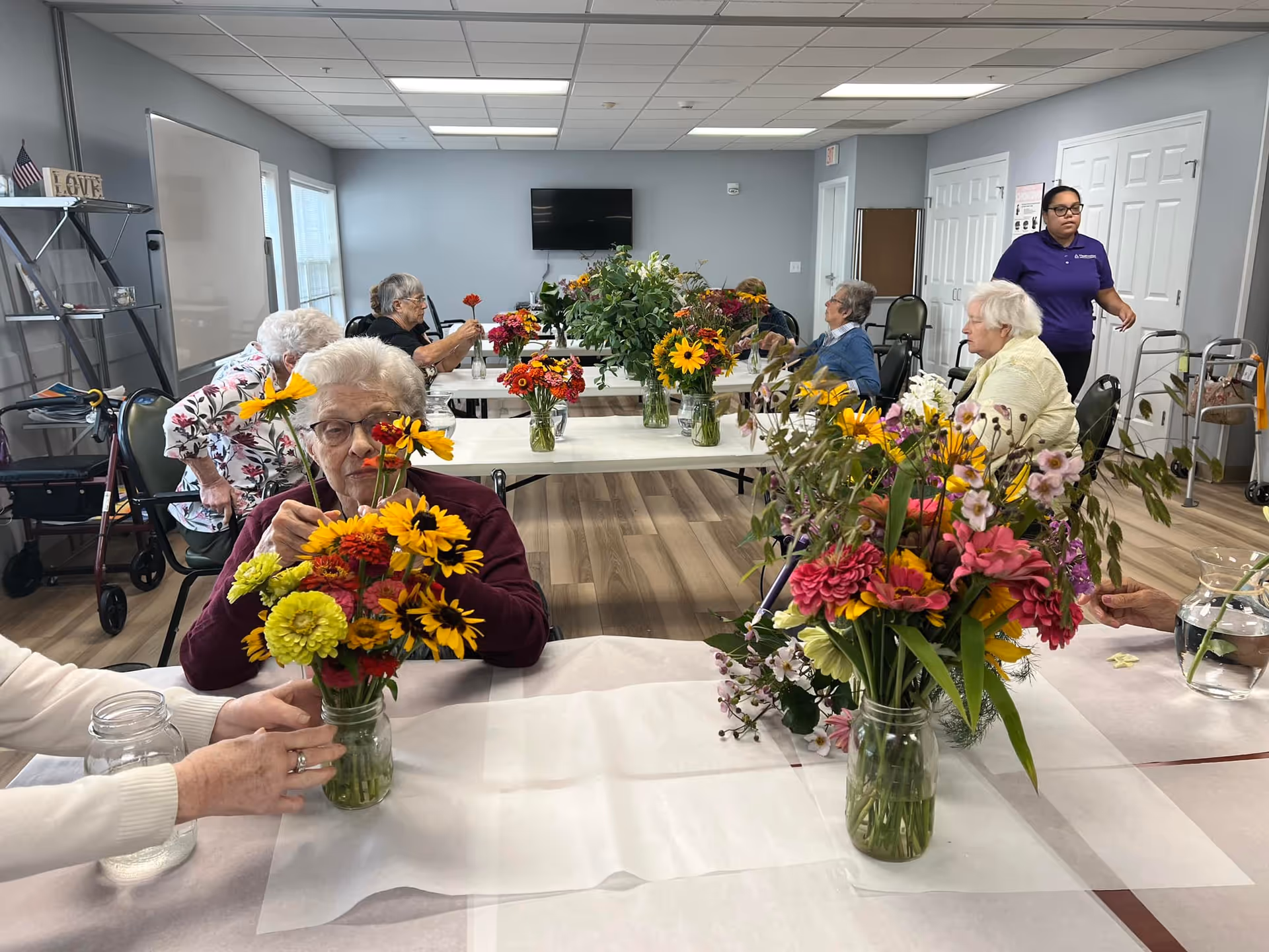 A group of elderly individuals seated at tables in a bright room, arranging colorful flower bouquets in glass jars. A caregiver in a purple shirt stands nearby. The room has light gray walls, wood flooring, and a television mounted on the wall.