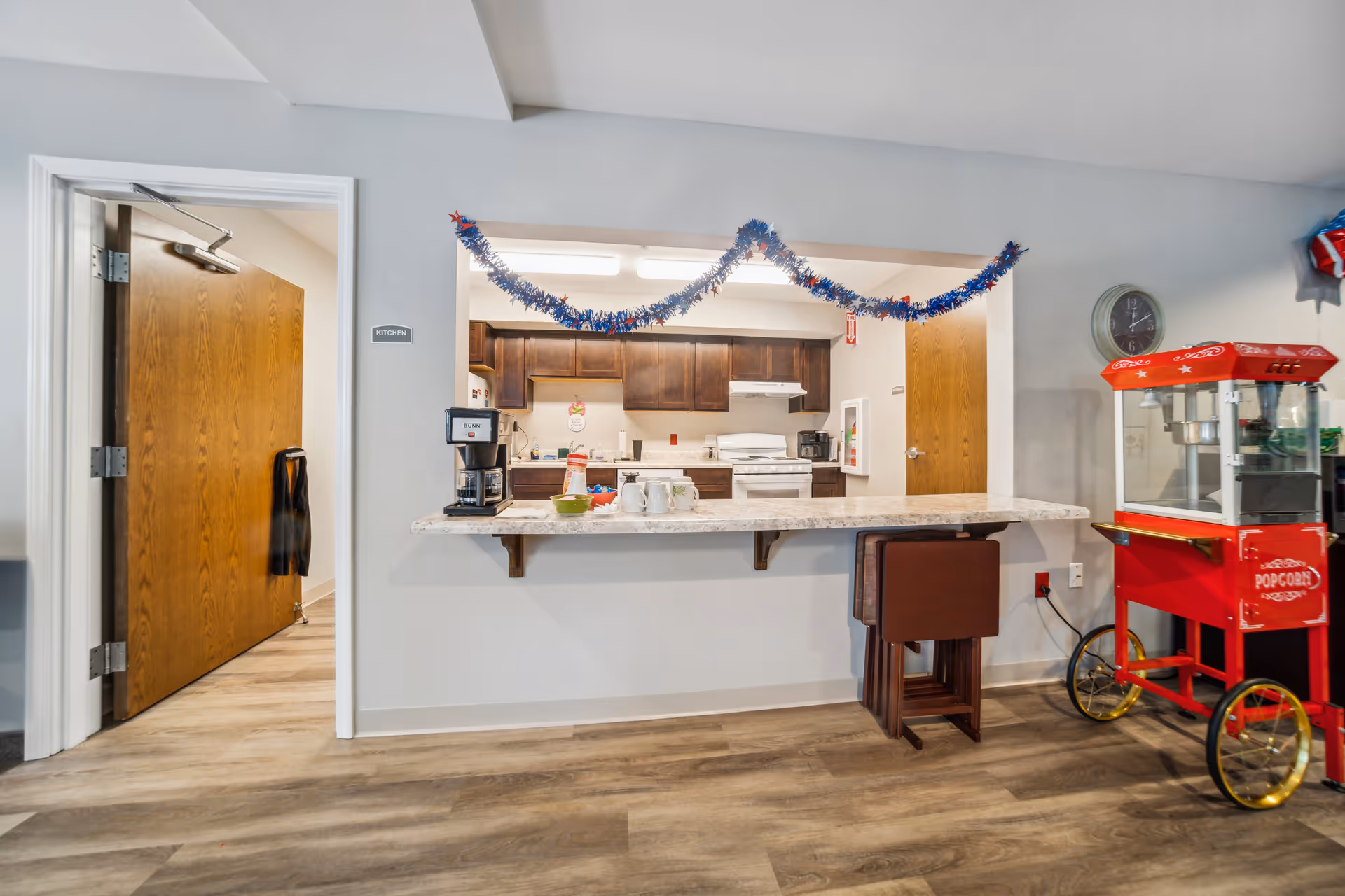 Interior view of a kitchen area in a senior living facility with a countertop bar, coffee maker, mugs, and bowls. The kitchen has wooden cabinets, a stove, and a refrigerator. There is a red popcorn machine on wheels to the right and a clock on the wall. Blue and red garland decorations hang above the kitchen pass-through window. A door labeled 'Kitchen' is open to the left.
