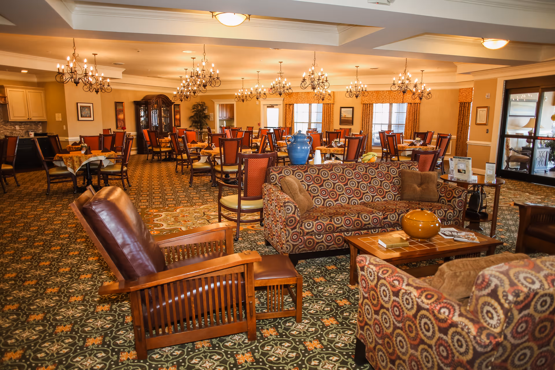 A spacious and warmly lit dining room with multiple tables and chairs arranged for meals. The room features patterned carpet, chandeliers hanging from the ceiling, and a sitting area with patterned sofas and wooden armchairs in the foreground. Large windows with curtains and a glass door leading outside are visible in the background.