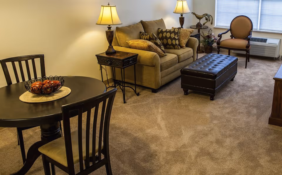Cozy living area with a beige sofa, tufted leather ottoman, round dining table with chairs, table lamps, and carpeted floor.