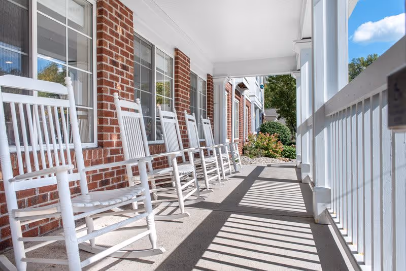 A row of white wooden rocking chairs lined up on a covered porch with a brick wall and windows on one side and white railing on the other. The porch overlooks a garden area with bushes and trees under a blue sky with some clouds.