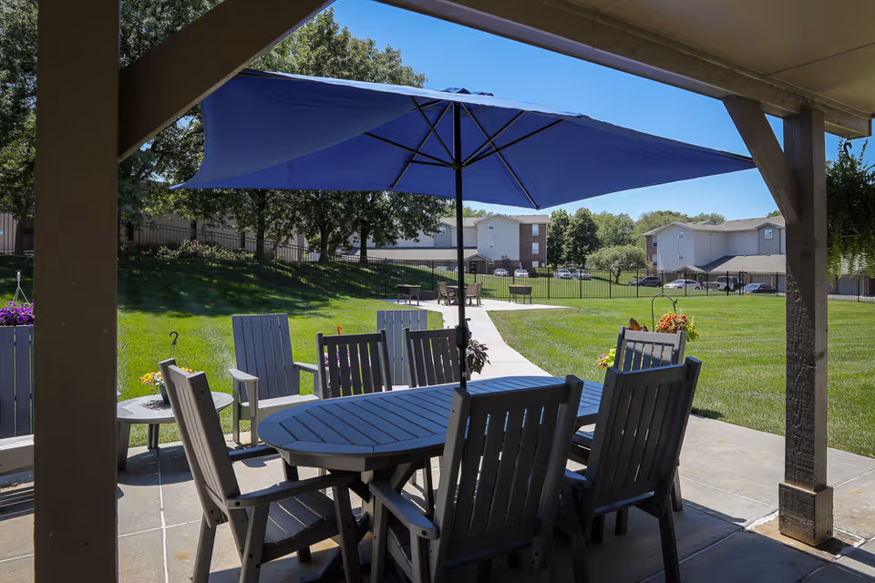 Shaded patio with a table, chairs, and a blue umbrella overlooking a grassy courtyard and nearby buildings.