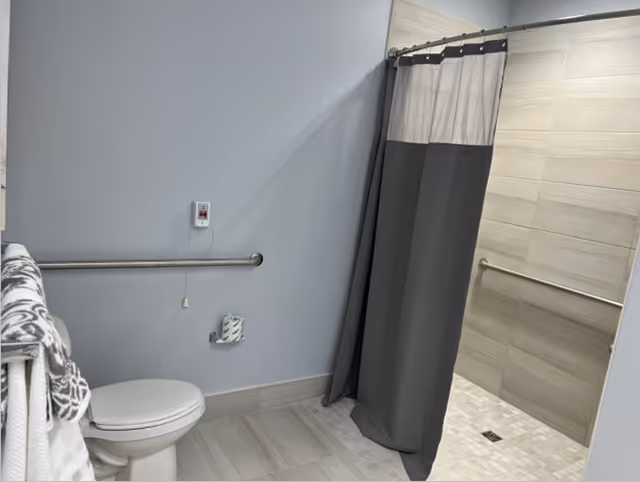A bathroom with a toilet, a grab bar on the wall, a towel rack with folded towels, and a walk-in shower with a gray and white shower curtain and tiled walls and floor.