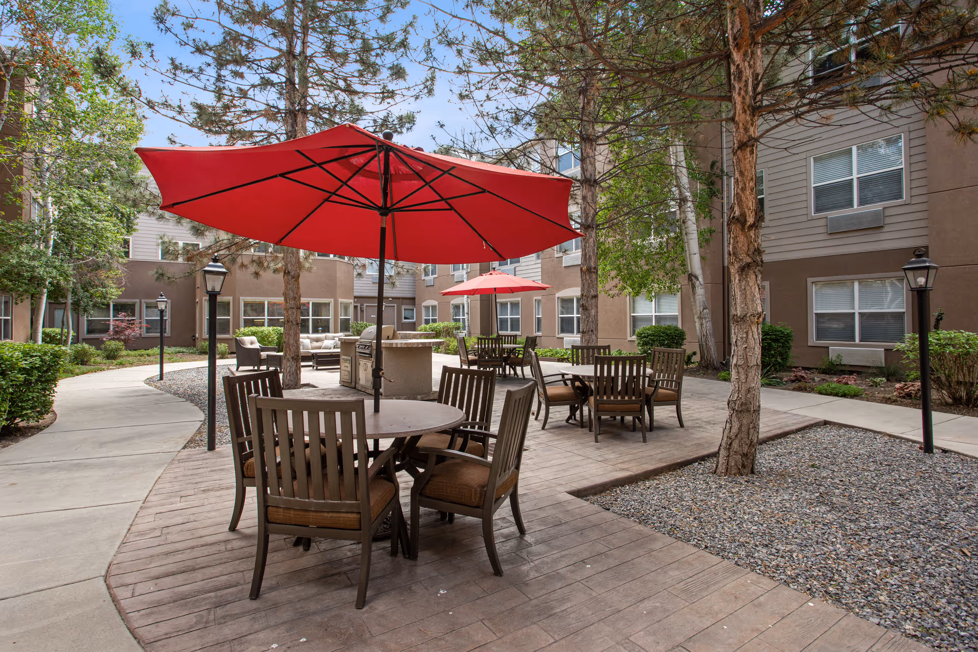 Outdoor courtyard area at Valencia at Cottonwood Heights featuring round wooden tables with chairs and large red umbrellas. The space is surrounded by trees, shrubs, and multi-story residential buildings with windows. There are paved walkways and a gravel section adjacent to the seating area.
