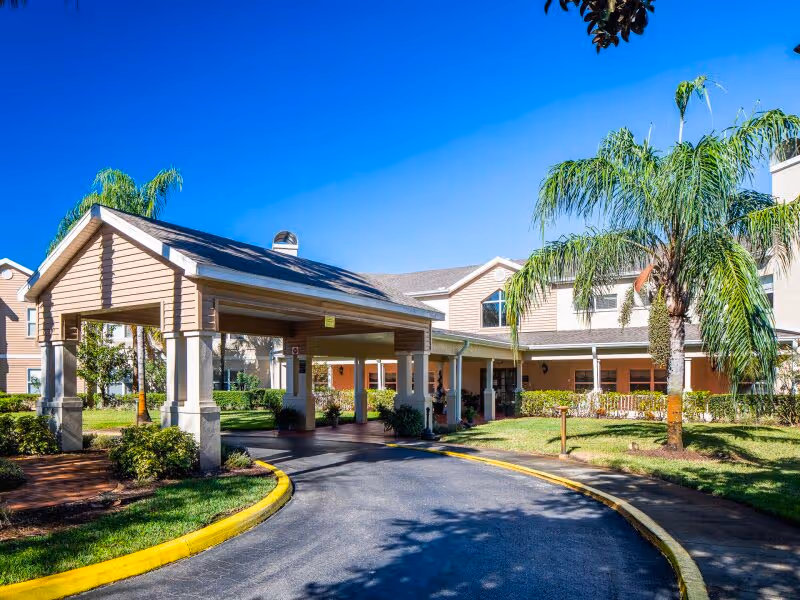 Exterior view of Chapman Manor at Lutz showing the entrance driveway with a covered drop-off area, surrounded by palm trees and landscaping under a clear blue sky.
