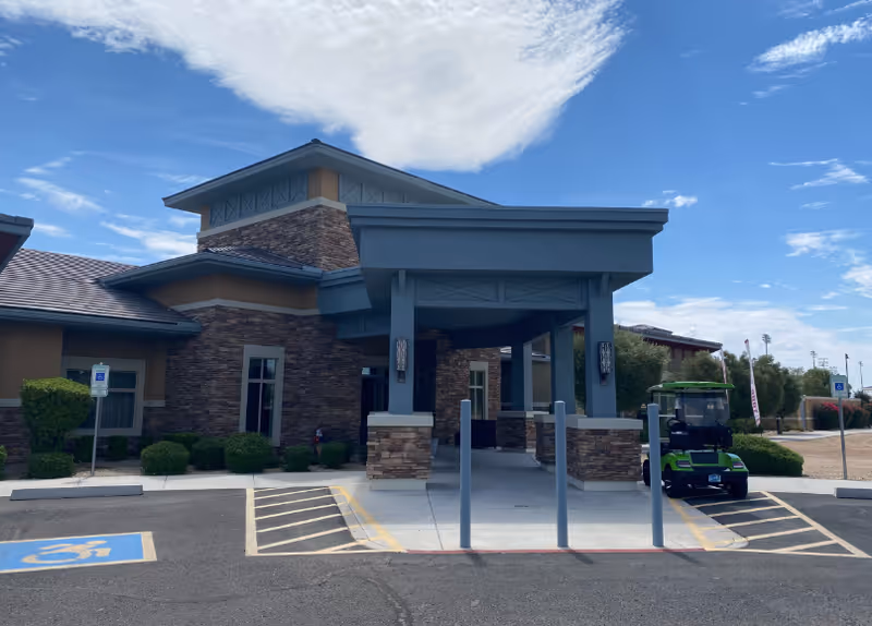 Front entrance of a single-story brick-and-stucco memory care facility with a covered porte-cochère, accessible parking spaces, and a small green cart parked to the right.