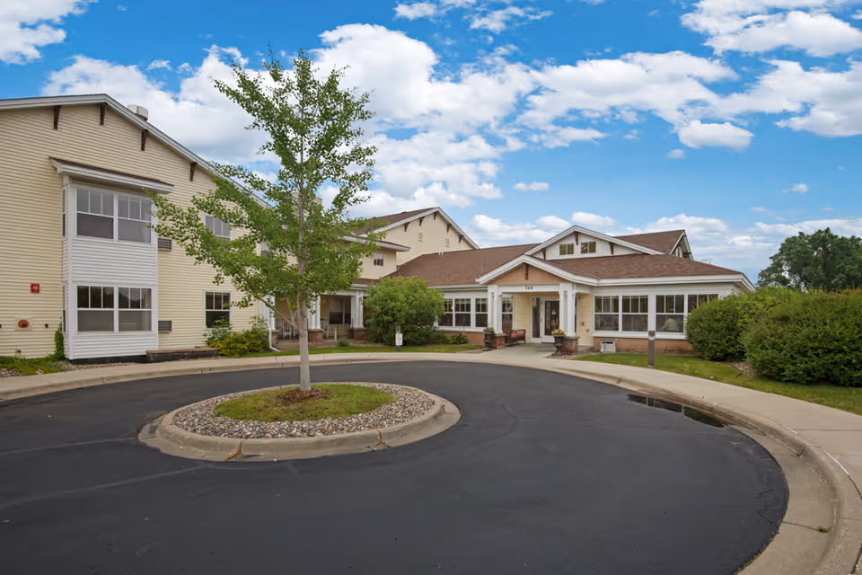 Exterior view of a senior living facility building with beige siding, multiple windows, and a brown roof. There is a circular driveway with a small tree and landscaping in the center. The sky is partly cloudy with blue patches.