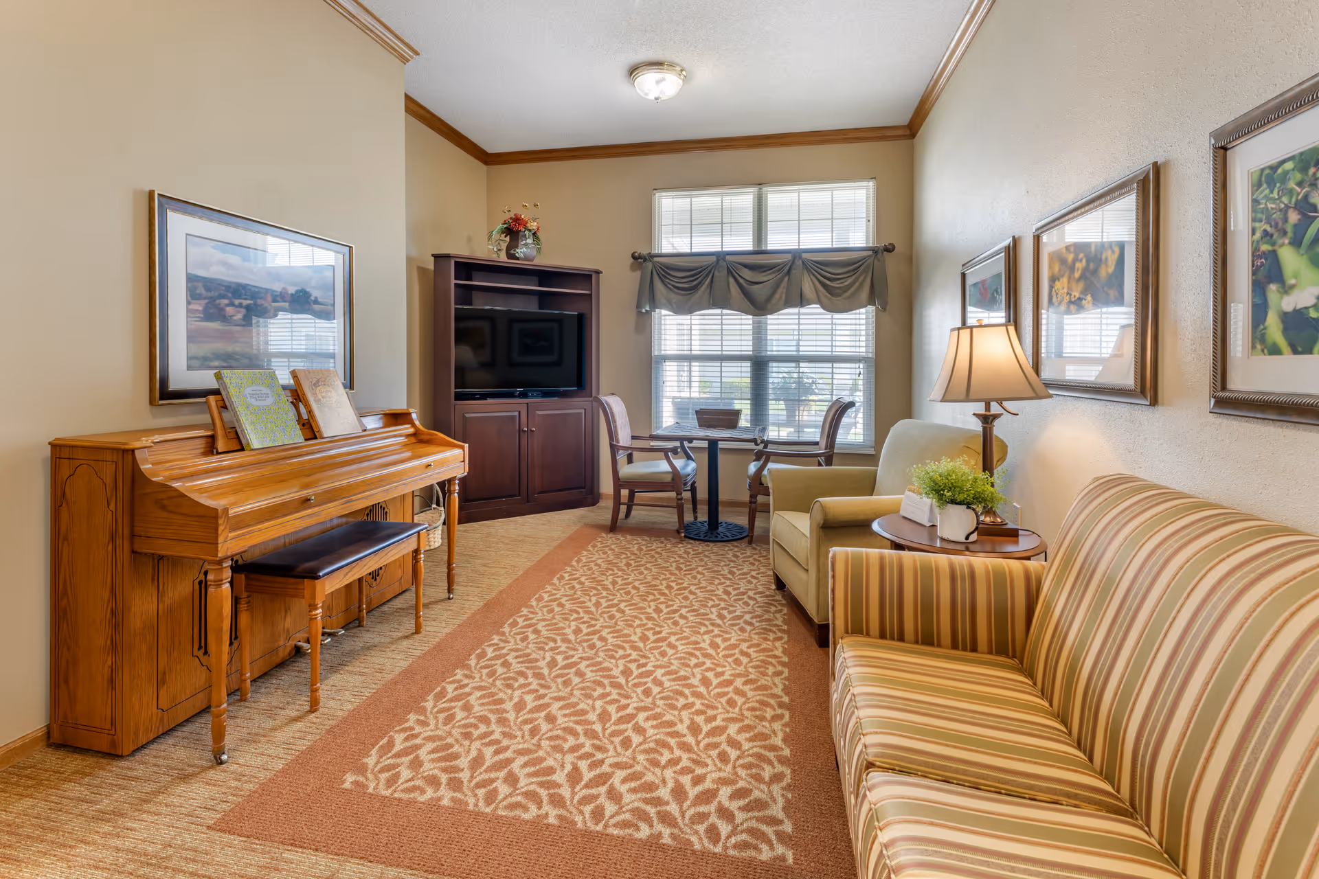 A cozy living room area with a striped sofa on the right, a small round table with a lamp and plant, an armchair, a small dining table with two chairs near a window with blinds and a valance, a wooden piano with a bench and sheet music on the left, and a dark wood TV cabinet with a flat-screen TV. The walls are decorated with framed artwork and the floor is carpeted with a patterned rug.