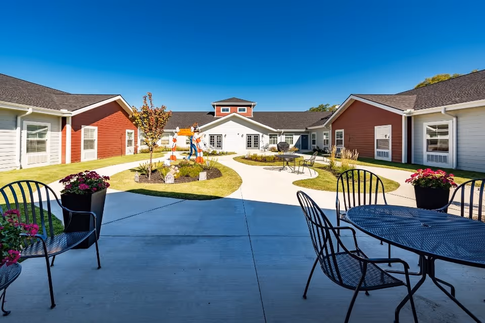 Outdoor courtyard area of a senior living facility with a clear blue sky, featuring a concrete walkway, metal patio tables and chairs, potted flowers, small landscaped garden beds, and a colorful scarecrow decoration. The surrounding buildings have red and white siding with multiple windows.