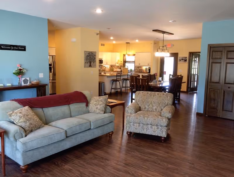 Open living area with a sofa and armchair in the foreground and a dining table and kitchen visible in the background.