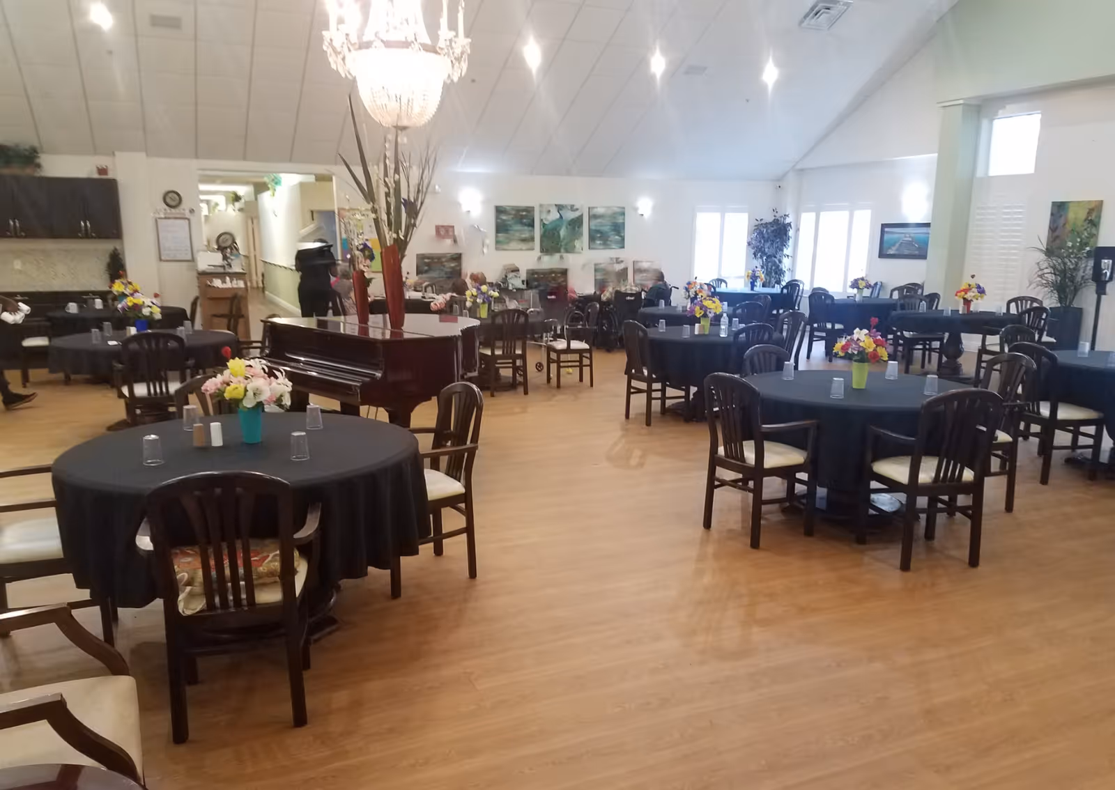 A spacious dining room with multiple round tables covered with black tablecloths, each adorned with colorful flower arrangements. Wooden chairs with cushions surround the tables. A grand piano is positioned near the center of the room under a large chandelier. The room has wooden flooring, light-colored walls, and several paintings and plants along the walls. There are a few people in the background, some seated and some standing.