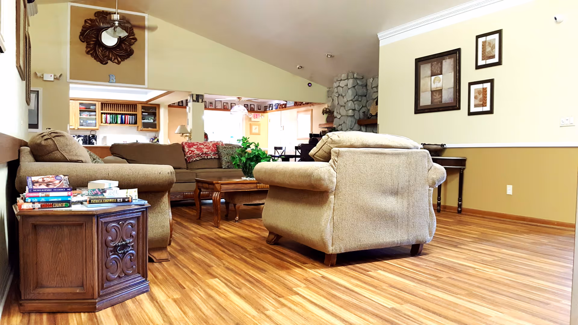 A cozy living room area with beige upholstered armchairs and a sofa arranged around a wooden coffee table. The room has wood flooring, light yellow walls with framed artwork, and a stone fireplace in the background. A small wooden side table next to the armchair holds a stack of books. The kitchen area is visible in the background with wooden cabinets and colorful plates on display.