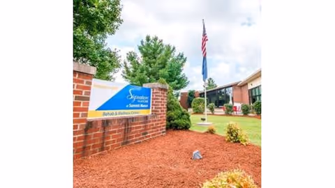 Outdoor view of Signature HealthCARE at Summit Manor Rehab & Wellness Center showing a brick sign with the facility name, a flagpole with an American flag, and a well-maintained lawn with bushes and trees under a partly cloudy sky.