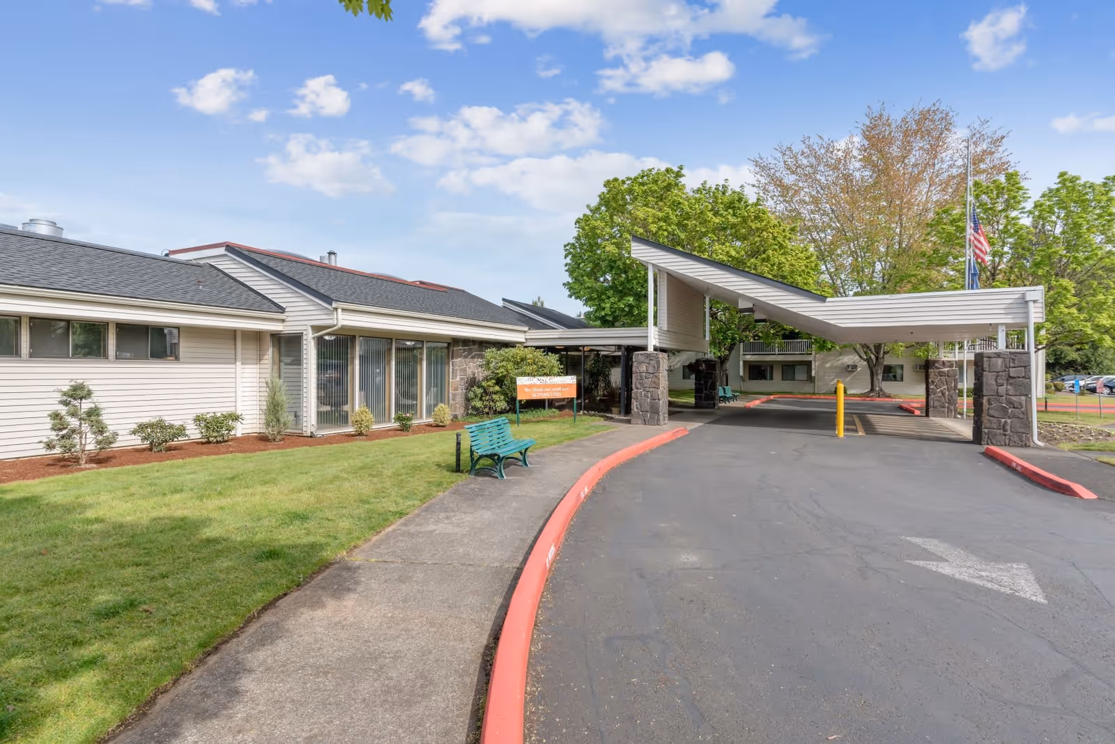 Exterior view of Summerfield Estates Retirement Community showing a covered entrance with stone pillars, a driveway with a red curb, a green bench on a sidewalk, and a building with large windows surrounded by green trees and a partly cloudy sky.