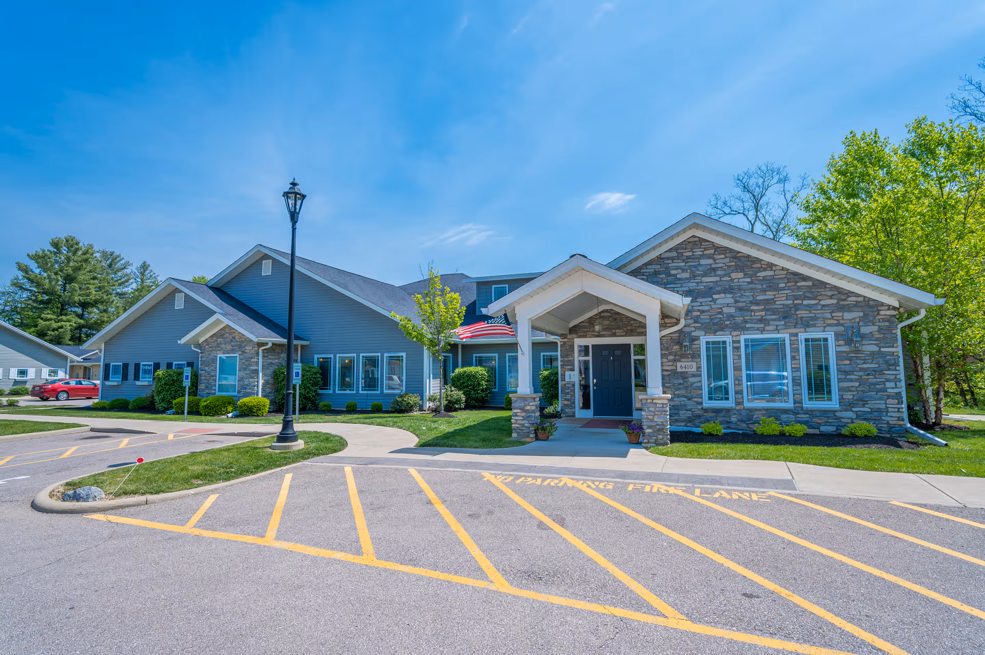 Exterior view of a single-story senior living facility building with stone and blue siding, a covered entrance with pillars, an American flag, and a parking lot with yellow no parking fire lane markings. Trees and greenery surround the building under a clear blue sky.